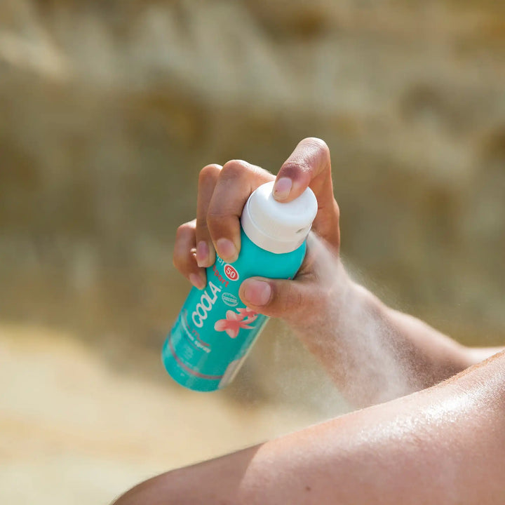 Teal-colored bottle of Coola sunscreen being held in a hand.