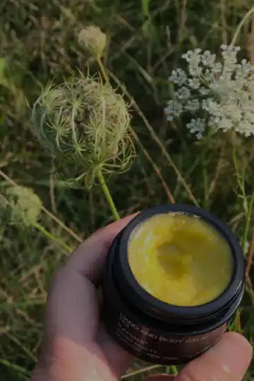 Yellow cream or balm in a dark jar being held against a natural backdrop.